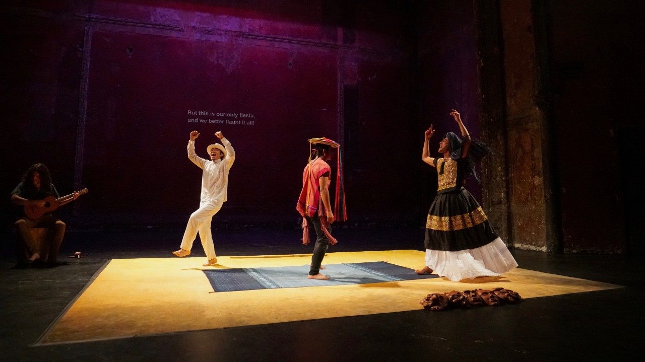  The cast of "Andares" dance and perform on stage. They stand on a yellow rectangle, in the center of which is a small black and white woven rug. To the far left, a musician sits on a wooden box and plays an acoustic guitar. Behind the actors, the English translation is projected onto a screen. The actor on the left wears a white shirt, pants, and hat, left leg lifted slightly in front of him, both arms raised above his head. In the middle, a brown person wears a red shirt, black pants, and a straw hat with multicolored ribbons cascading down from the rim. On the right, a brown woman wears a black, gold, and white traditional dress and lifts both arms above their head.