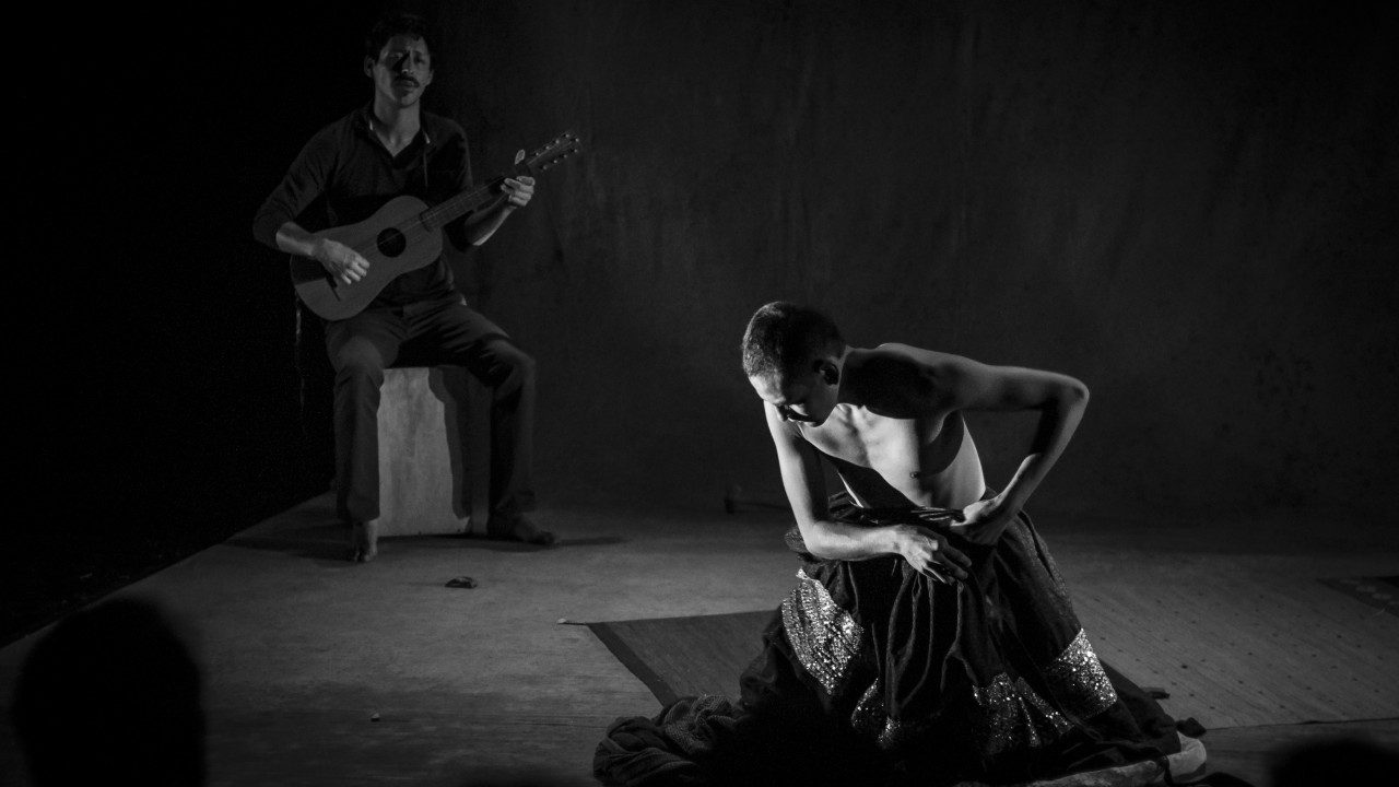  An actor of "Andares" kneels on the floor on a woven rug. They have a closely shaved head and are fastening the waist of a long, full skirt with a band of sequins. Behind them, a musician sits on a wooden box and plays and acoustic guitar in this black and white image.