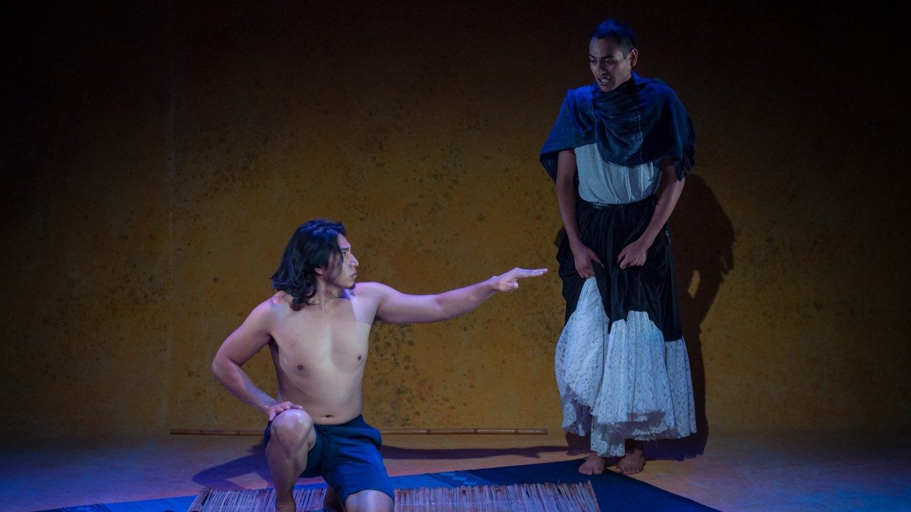  Two of the actors in "Andares" perform the play. On the left, a brown man with long dark brown hair and grey shorts kneels on a woven rug, his left arm extended sideways at the shoulder. On the right is a person with a shaved head wearing a long, full black and white skirt, white shirt, and grey shawl over their shoulders.