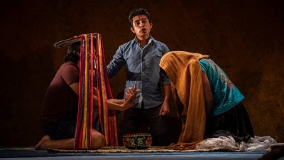  Actors from "Andares" kneel on a woven rug and perform. On the left, a person in a maroon shirt and dark shorts wears a round straw hat, multicolored ribbons streaming down from it, the ends tucked under their knees. On the right a person in a turquoise shirt and black and white skirt bends forward slightly, as if praying, with a mustard-colored shawl draped over their head. In the middle, a brown man with short dark brown hair wears a blue button down shirt and sings or speaks, looking towards the camera.