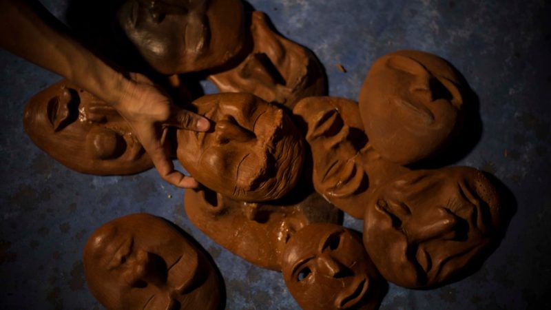  A pile of golden brown clay masks is on the grey floor. Someone reaches in, with one of the masks in their hand. Their skin is the same color as the masks.