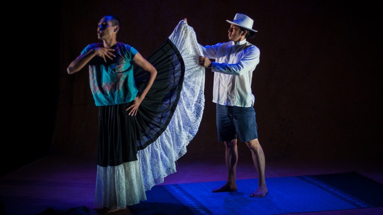  Cast members of "Andares" perform the play. On the right is a brown man in a white shirt and white hat, and dark blue shorts. He holds up the skirt of the person on the left, who has a closely shaved head and wears a turquoise shirt. The skirt is long, full, and black and white.