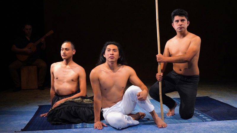  The cast of "Andares," three brown men, sits on a woven rug, all bare-chested. The actor on the left has a shaved head and wears a long, full skirt. The actor in the middle has long dark brown hair and wears white pants. The actor on the right has short dark brown hair, wears black pants, and holds a wooden walking stick vertically against the ground in front of him.