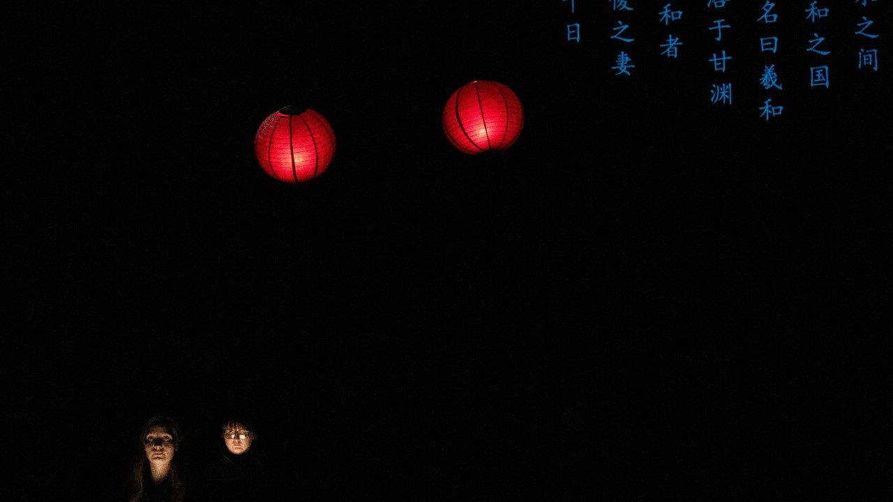  A production shot from "Book of Mountains and Seas." In the bottom left corner are the uplit faces of two vocalists. In the middle, towards the top, are two red round paper lanterns. In the top right corner, blue Chinese characters are projected onto a screen. All the rest is dark.