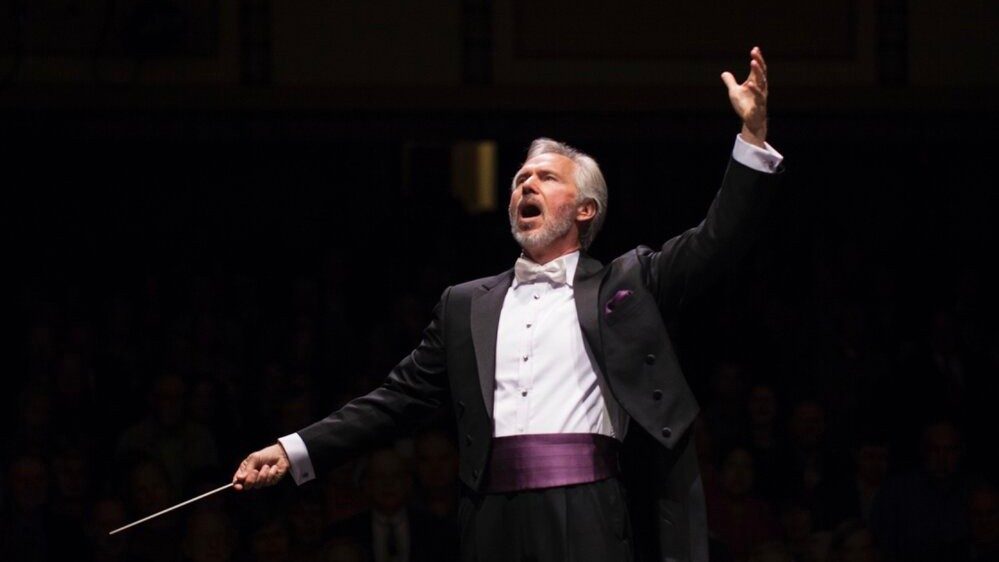  Steven White, conductor and artistic director of Opera Roanoke, is a white man with grey hair. He wears a black suit, white button down shirt, and eggplant cummerbund and pocket square, his arms thrown wide energetically, a conducting baton in his right hand.