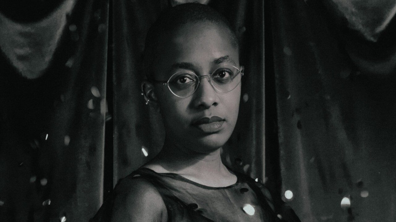  Cécile McLorin Salvant, a light-skinned Black woman with a closely shaved head and round thin wire-framed glasses, wears a sheer dress in front of a fabric backdrop in this black and white image.