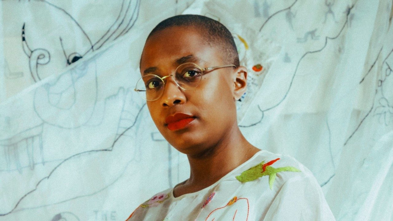  Cécile McLorin Salvant, a light-skinned Black woman with a closely shaved head and round thin wire-framed glasses, wears a matte red lipstick and a white shirt embroidered with brightly colored cartoonish figures. Behind her is an offwhite fabric backdrop, onto which illustrations have been drawn in black marker.