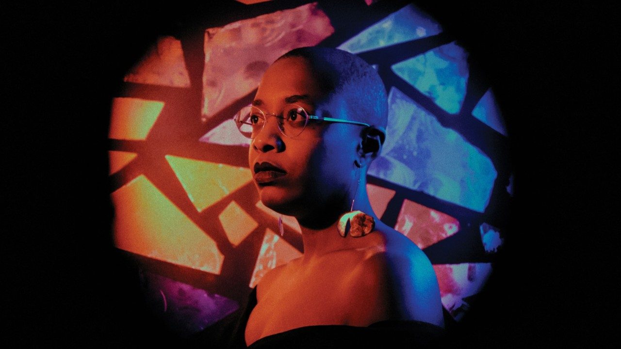 Cécile McLorin Salvant, a light-skinned Black woman with a closely shaved head and round thin wire-framed glasses, wears a dark lipstick and an off-the-shoulder black dress and large statement earrings. She is lit in and orangey-pink light, and behind her is a round projection of shards of glass or ceramic in shades of orange, blue, pink, red, and purple.