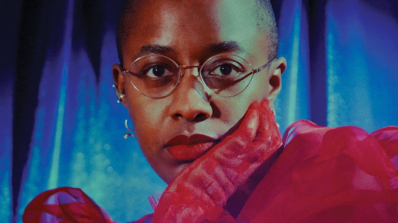 Cécile McLorin Salvant, a light skinned Black woman with round thin wire-framed glasses, wears a matte red lipstick, flouncy red dress, and red mesh gloves. She cradles her chin in her left hand and looks directly at the camera. Behind her is a blue fabric backdrop.
