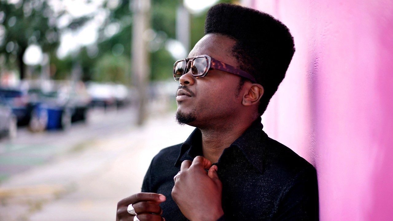  Musician Cimafunk, a Black man with a high top, wears a black button down shirt and sunglasses, in front of a pink exterior wall of a city building. His hands are at his collar as if he's straightening a tie.