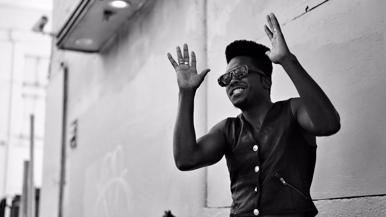  Musician Cimafunk, a Black man with a high top, wears a black button down vest and sunglasses, and throws his hands up near his head in front of the exterior wall of a city building in this black and white image.