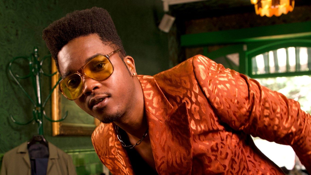  Musician Cimafunk, a Black man with a high top, wears an orange satin leopard print blazer and transparent amber-colored sunglasses. He leans towards his right while looking at the camera. He's inside a bar painted a lush forest green, large picture window in the background behind him.