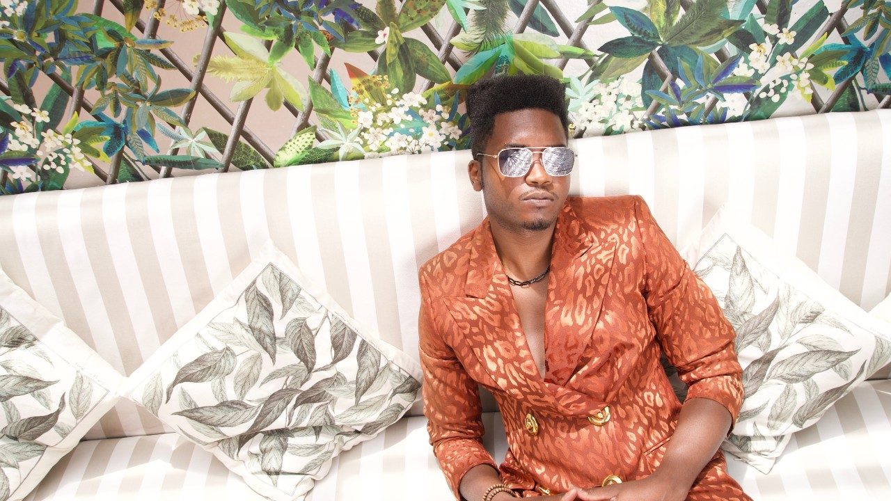  Musician Cimafunk, a Black man with a high top, wears an orange satin leopard print double breasted blazer over his bare chest and reflective aviator sunglasses. He sits on a beige and white striped upholsered couch with floral-patterned throw pillows. The wallpaper behind him looks like colorful vines and flowers growing up a chainlink fence.