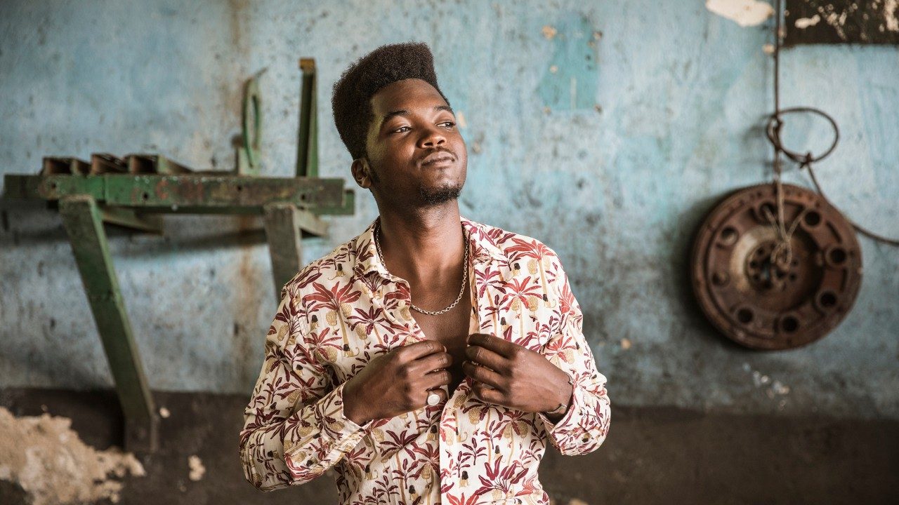  Musician Cimafunk, a Black man with a high top, wears a floral patterned button down shirt, and he appears to be closing the top button. He's in a rough-looking garage, and he looks slightly to the left of the camera.