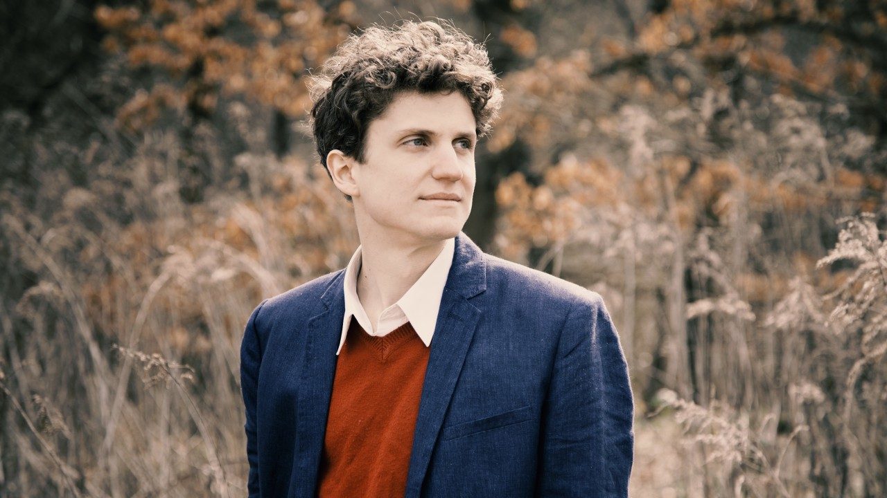  Zoltan Fejérvári, the pianist who will perform with Concerto Budapest Symphony Orchestra, stands in front of tall grasses. He is a white man with curly brown hair wearing a white button down shirt, red sweater, and blue blazer.