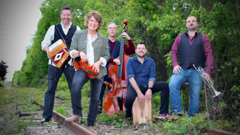  Eileen Ivers and her band, Universal Roots, pose on an abandoned railroad track. Leafy green trees are visible behind them. The musicians hold their instruments, from left, an accordion, fiddle, bass, and trumpet. The drummer sits on a wooden box.