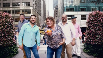  Fiddler Eileen Ivers, a white woman with short layered blonde hair, wears a blue paisley patterned shirt and jeans and holds an apple towards the camera. Her band surrounds her, two men on each side, as well as two bushes with pink flowers. They appear to be in New York City.
