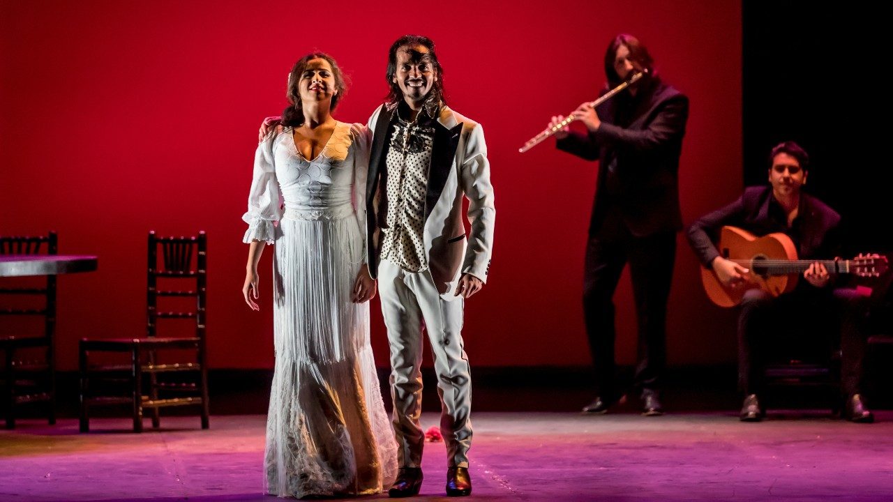  Flamenco dancer Farruquito, on the right, and a female flamenco dancer wear all white and stand next to each other on stage while two musicians play in the background.