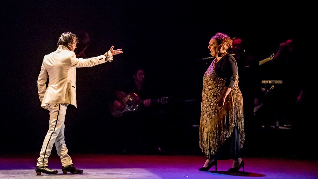 Flamenco dancer Farruquito, on the left, wears a white suit and black shoes on stage. He extends his right arm towards a middle aged woman in a long brown dress, who sings.