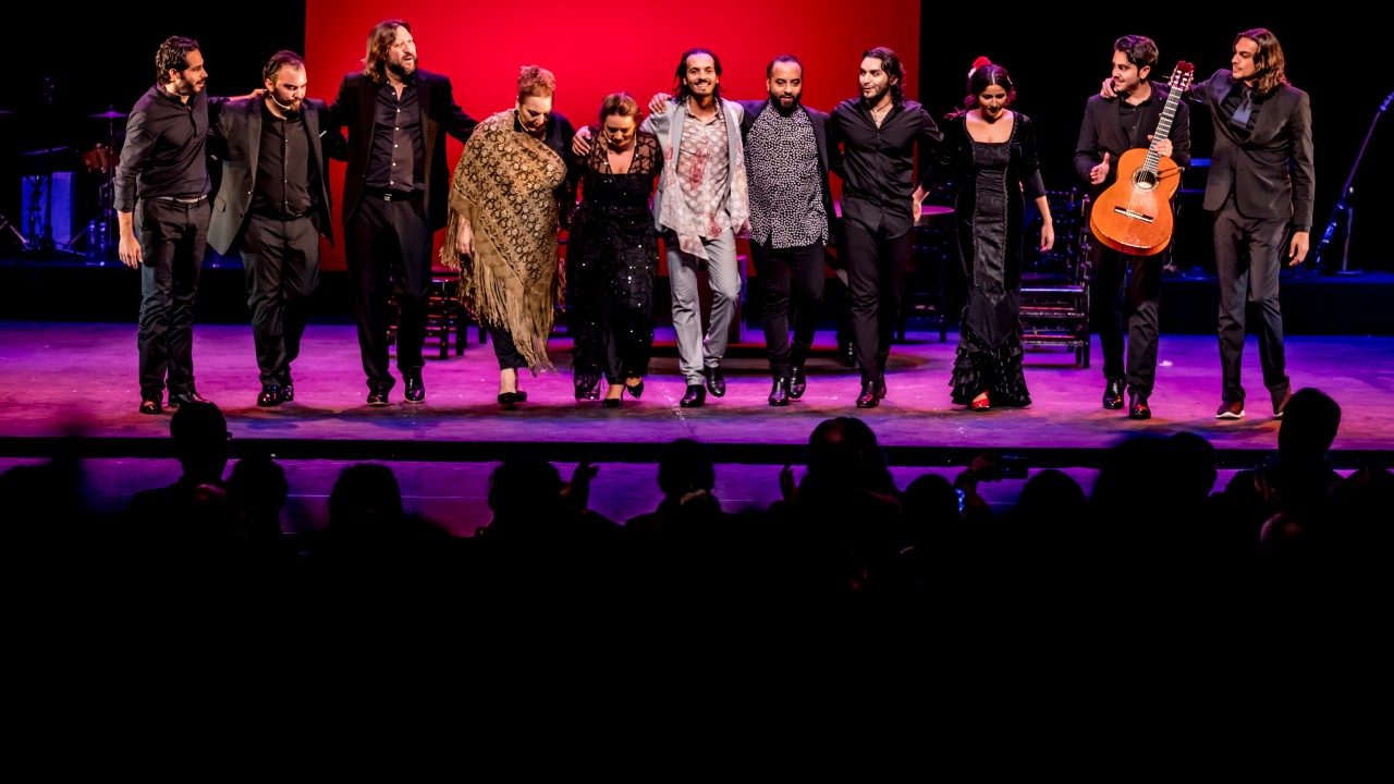  Dancers, singers, and musicians with Farruquito stand in a line on stage at during a performance while the audience claps for them.