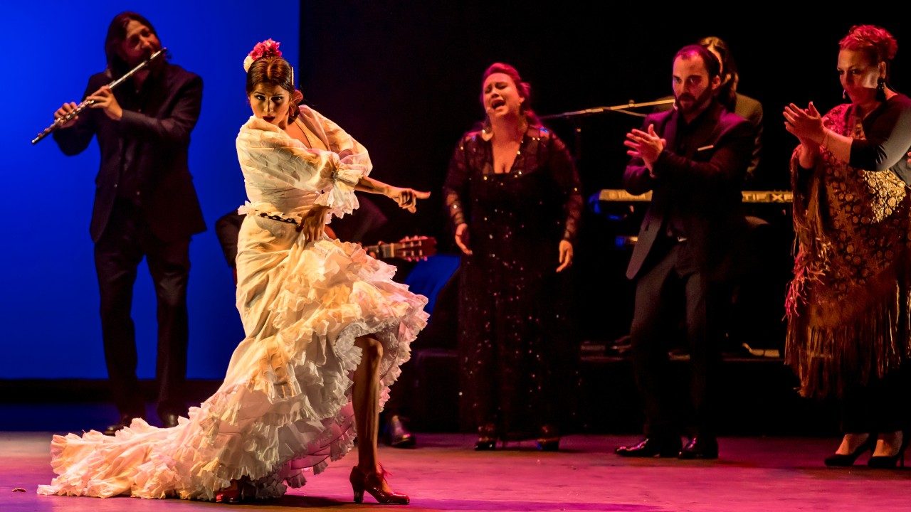  A female flamenco dancer in a long white dress with tiered ruffles around the hem dances on stage, while musicians and singers perform behind her.