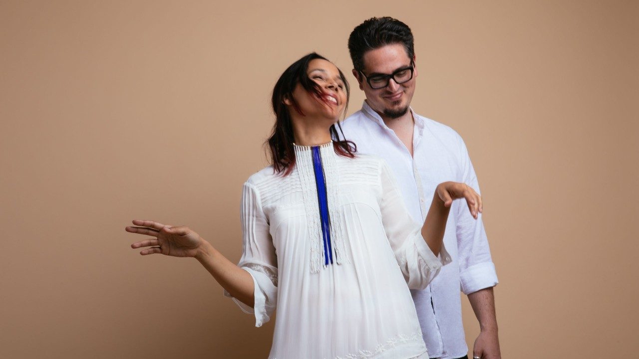  Rhiannon Giddens, at left, a brown-skinned woman with medium length brown hair, wears a white dress and a white and blue thread necklace. She appears to twirl or move to music, a gentle happy smile on her face and both arms bent at the elbows. Francesco Turrisi, at right, a white man with dark brown hair and goatee and black framed glasses, smiles and gazes down towards the ground. He wears white button down shirt, and both stand in front of a brown background.