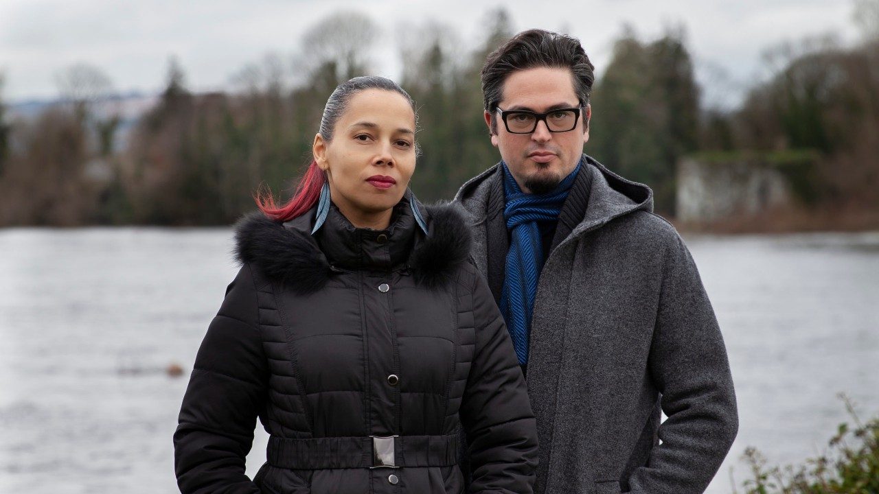  Rhiannon Giddens, left, is a light skinned Black woman with brown and red hair slicked back into a ponytail. She wears feather earrings, a heavy black winter coat, and wine-colored lipstick. On the right is Francesco Turrisi, a white man with medium length dark brown hair, goatee, and dark framed square glasses. He wears a heavy grey wool winter coat and blue scarf. They stand in front of a river.