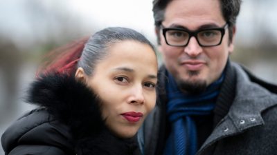  Rhiannon Giddens, left, is a light skinned Black woman with brown and red hair slicked back into a ponytail. She wears a heavy black winter coat and wine-colored lipstick. On the right is Francesco Turrisi, a white man with medium length dark brown hair, goatee, and dark framed square glasses. He wears a heavy grey wool winter coat and blue scarf. They stand in front of a river.