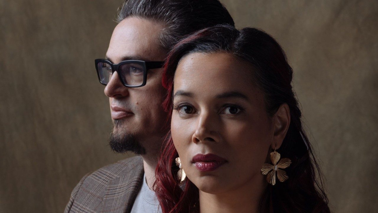  Francesco Turrisi (at left), a white man with dark brown hair and goatee and black framed glasses wears a brown plaid jacket over a grey T-shirt and looks towards his right. In front of him, Rhiannon Giddens (at right), looks towards the camera, her red-brown hair slightly curled and half up. She wears large gold flower earrings and a raspberry lipstick. They stand in front of a brown backdrop.