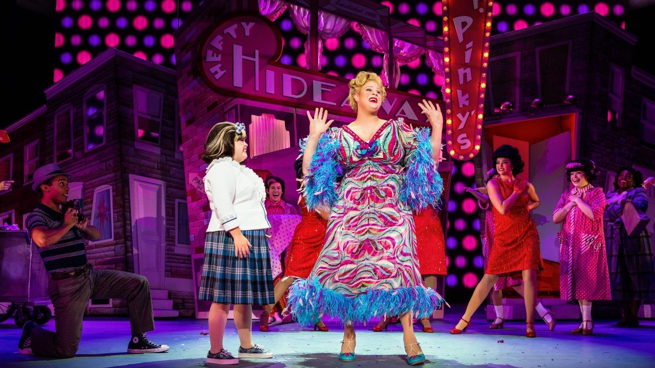  Cast members of "Hairspray" perform on a brightly colored, '60s-inspired set. In the foreground is a tall blonde person in a long multicolored patterned dress with blue feather trim at the hem and on the sleeves. Looking at her is a shorter heavyset girl with brown hair, a white button down shirt, and a plaid skirt.