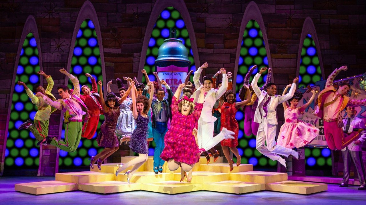  The cast of "Hairspray" sings and dances on set. Everyone jumps into the air with their arms extended above their heads in 60s-era costumes.