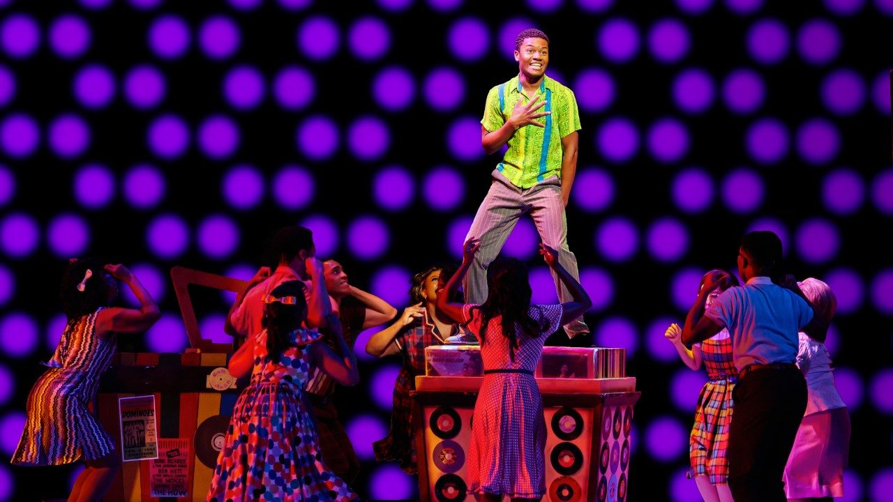  A Black man in a lime green and blue striped button down shirt and grey pants stands on a pedestal. Below him, other cast members dance in the shadows. The background is black with large purple polka dots in circular patterns.