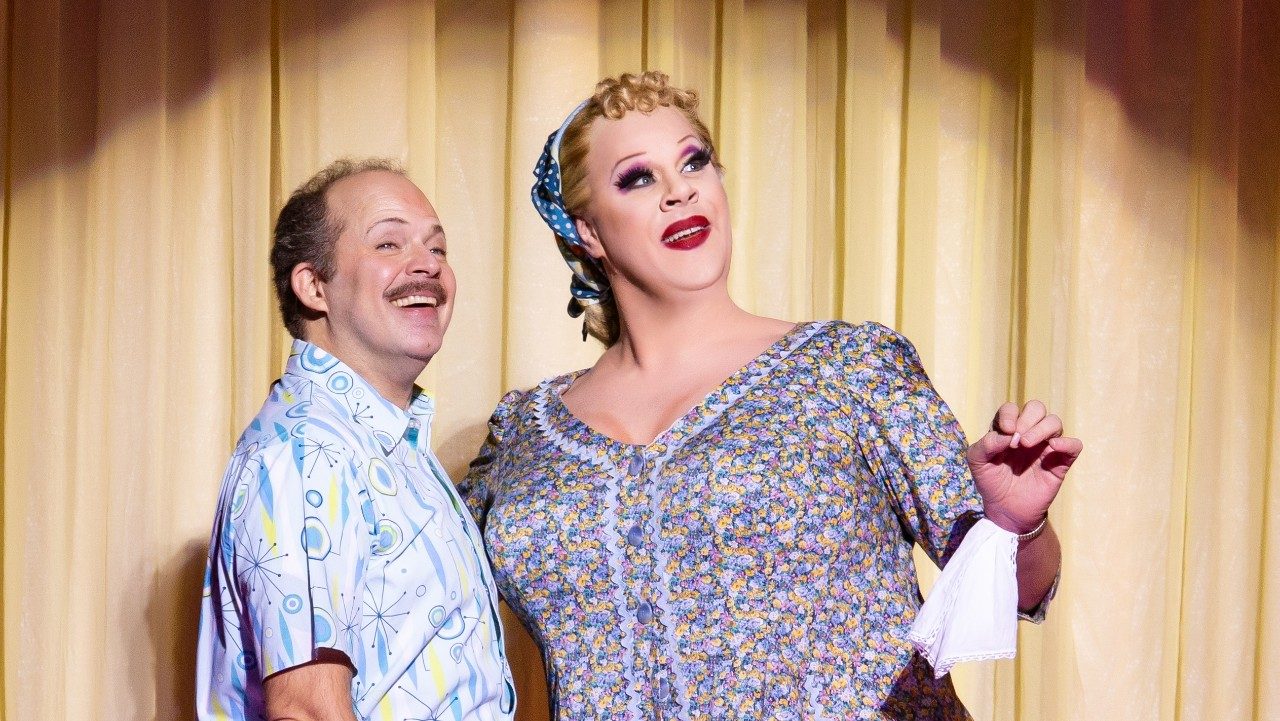  A white man in a patterned button down shirt stands with a white man in drag in front of a beige curtain. The man in drag wears a blonde curly wig under a blue handkerchief and a blue floral dress. Hanging from their wrist is a white handkerchief, and they wear a strong red lipstick.