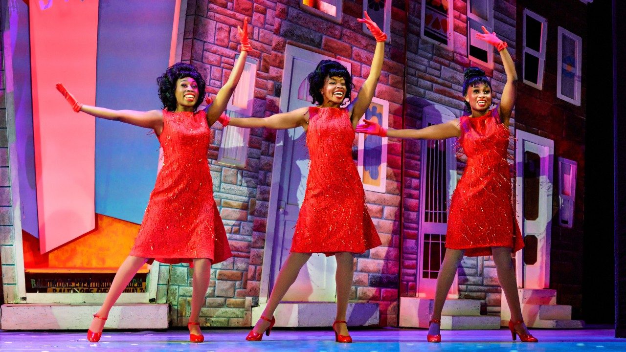  Three Black women in red A-line dresses, red gloves, and red high heels dance in front of a set that looks like stone apartment building fronts.