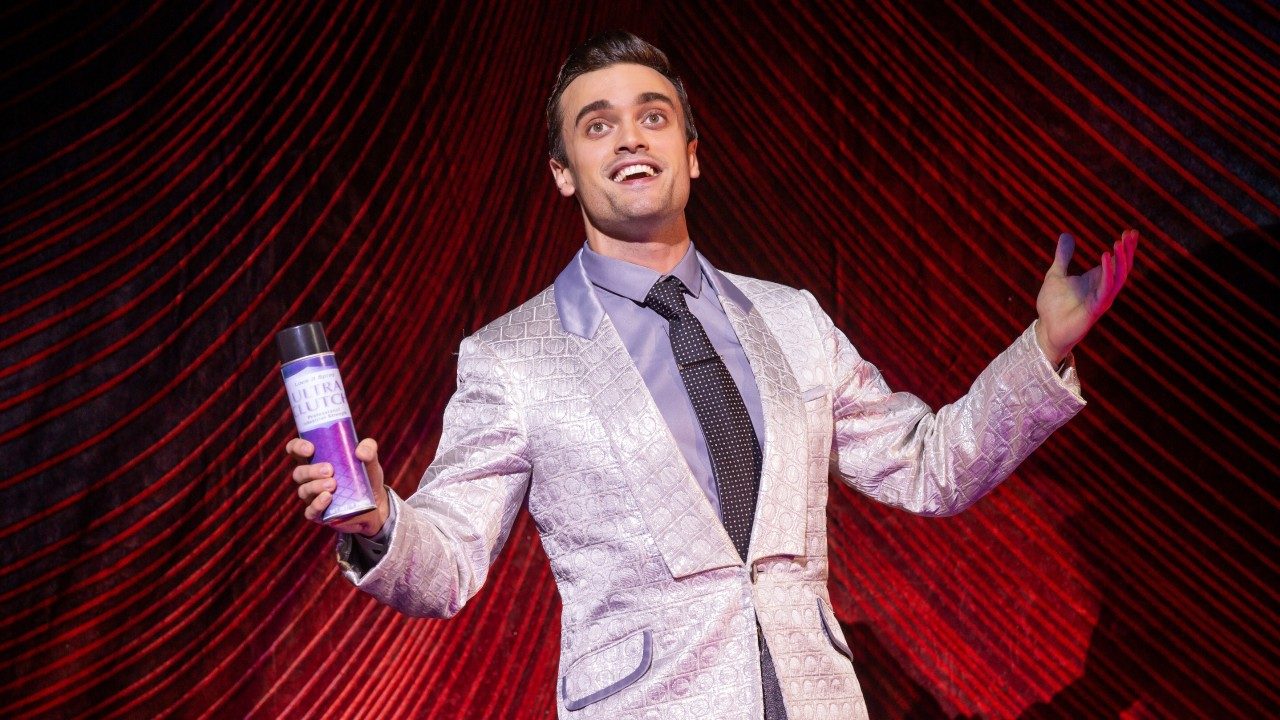  A white man with brown hair, a silver jacket, grey shirt, and dark grey tie holds a can of hairspray under the spotlight, arms outstretched in front of a red curtain. He looks like he's selling something.