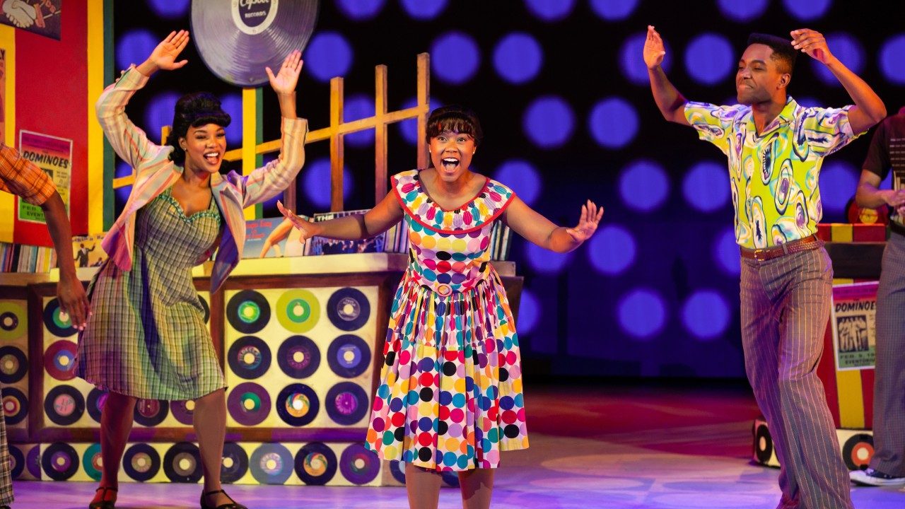  Two Black women and one Black man sing and dance on the brightly colored, '60s-inspired set for Hairspray.