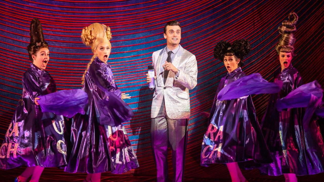  Four white women and one white man sing on stage. The women wear purple dresses and extravagantly styled hair. The man wears a silver jacket and pants, grey shirt, and dark grey tie.