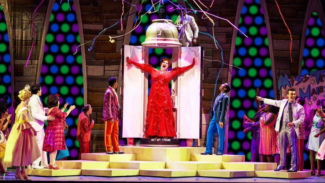  The cast of "Hairspray" stands on stage, some on yellow block stage risers, and look at a woman in a long red dress explode through two white doors, streamers released above her.