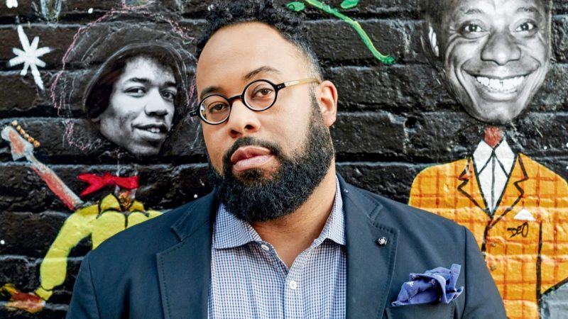  Poet Kevin Young, a Black man with short natural hair, a full and well-groomed beard, and small round glasses, stands in front of a mural on a brick wall featuring the faces of Bob Marley and James Baldwin. Young wears a blue plaid button down shirt, navy blue blazer, and a denim-blue pocket square.