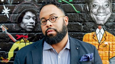  Poet Kevin Young, a Black man with short natural hair, a full and well-groomed beard, and small round glasses, stands in front of a mural on a brick wall featuring the faces of Bob Marley and James Baldwin. Young wears a blue plaid button down shirt, navy blue blazer, and a denim-blue pocket square.