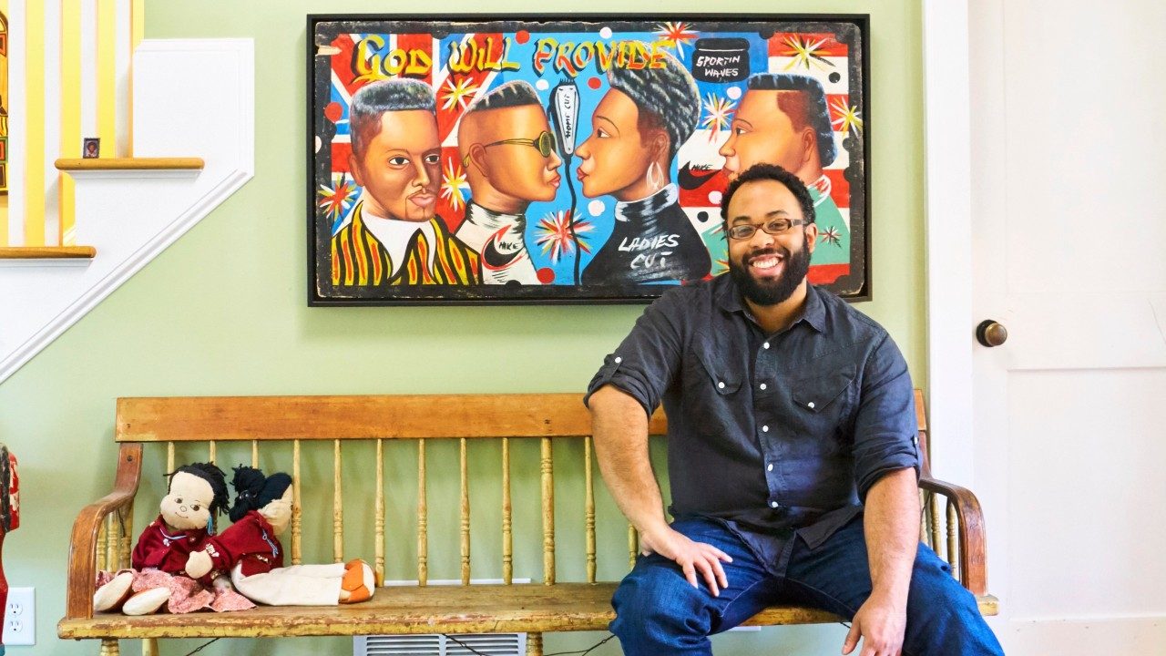  Poet Kevin Young, a Black man with short natural hair, a full and well-groomed beard, and dark framed glasses, sits on a wooden bench in his home. He wears a grey button down shirt and jeans. Two dolls sit on the other end of the bench, and behind Young, hung on a sage green wall, is a large art piece of four Black people with the words "God will provide."
