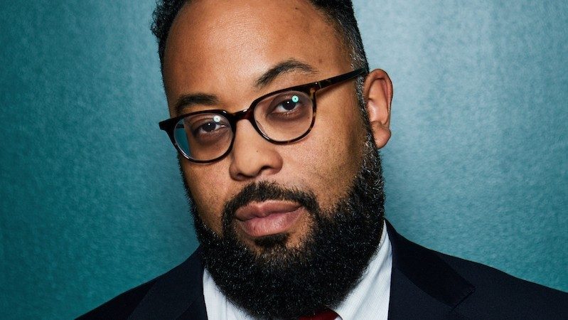  Poet Kevin Young, a Black man with short natural hair, a full and well-groomed beard, and thin tortoise shell framed glasses, stands in front of a teal wall. He hears a white button down shirt, tie, and black jacket.