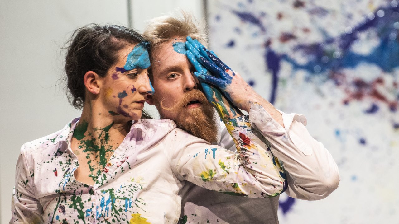  Two cast members of Machine de Cirque, a white woman with brown hair and a white man with blonde hair, blue eyes, and a blonde beard and curled moustache, wear white shirts covered in fresh paint splatters in front of a canvas. She stands in front of him to his right, looking towards his face, with her paint-covered left hand touching his forehead. He stands behind her and looks at the camera his paint-covered hand holding hers in place.