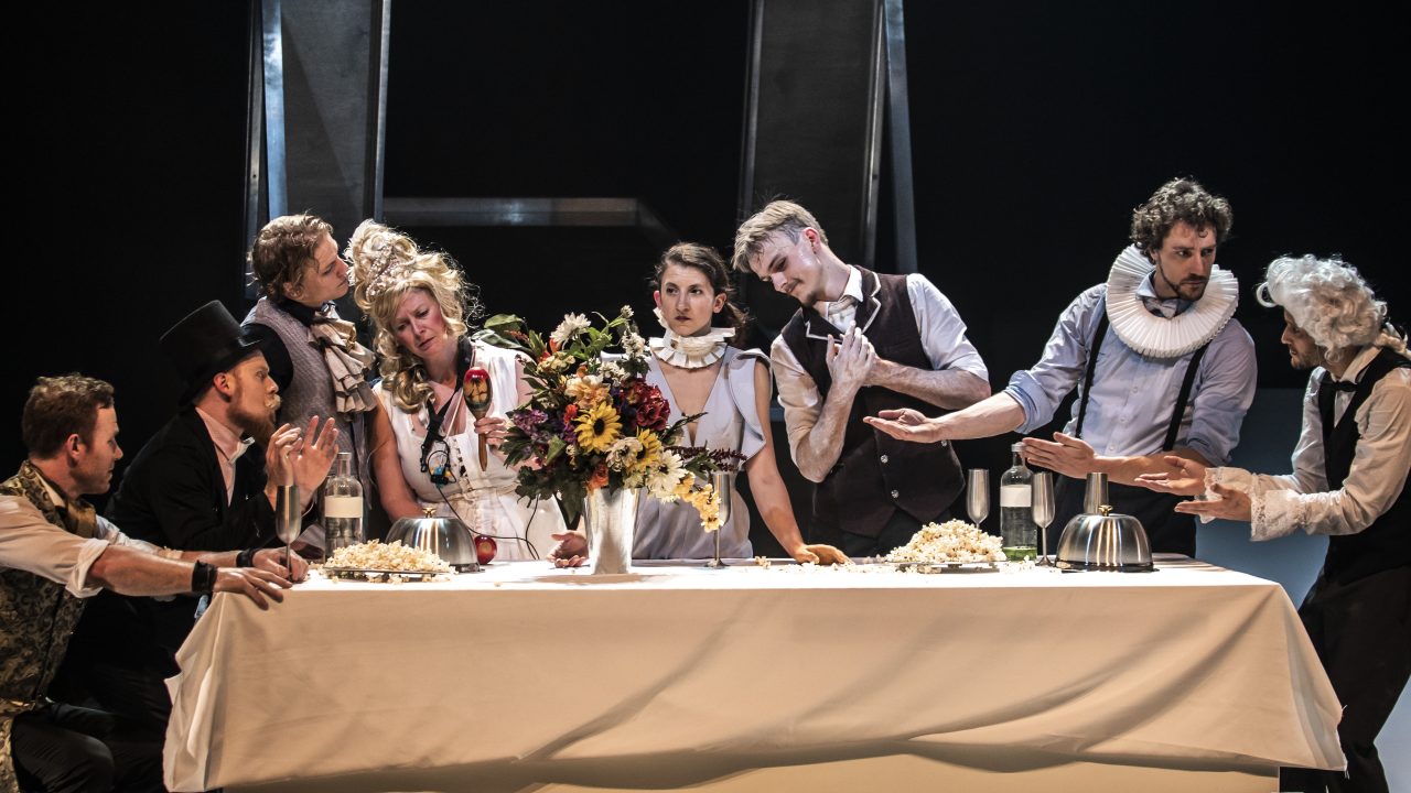  The cast of Machine de Cirque gather around a table draped in a white cloth, very similarly to "The Last Supper." The cast consists of six white men and two white women, the latter of whom are in the center of the group.
