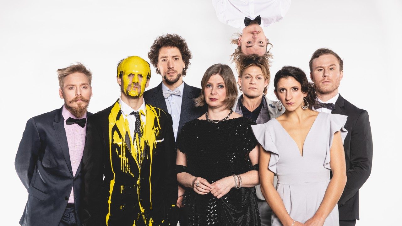  The cast of Machine de Cirque poses in front of a white background. The group is six white men and two white women. They all wear eveningwear. The second person from left, a white man, has had yellow paint dumped on top of his head. A white man appears to balance upside down on the head of the white man who is third from the right.