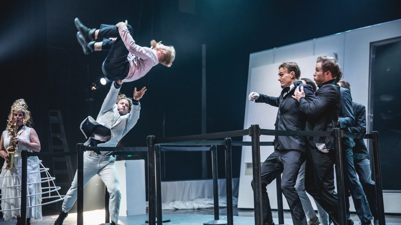  The cast of Machine de Cirque performs onstage. On the far left, a white woman with a tall blonde wig wears a caged skirt and plays a saxophone. In the center, a blonde man in a pink shirt and dark pants does a tucked knee backflip, while a man in a beige suit reaches out to catch or stop him. On the right, a cluster of four men and one woman bunch together as if trying to get out of the way of the flipping man.