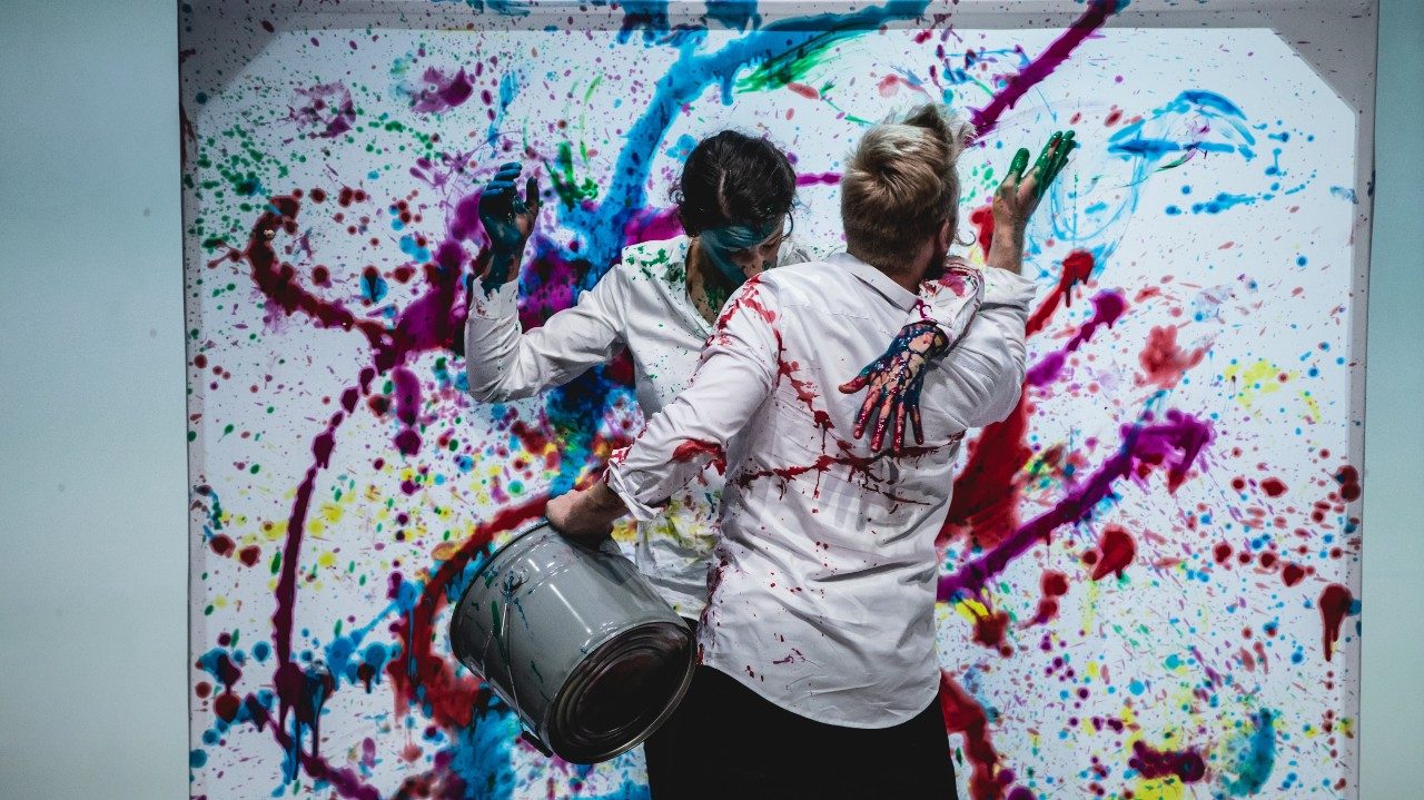  A white man with blonde hair and a white woman with brown hair splatter paint all over themselves and a large canvas. The woman has her back to the canvas facing the man, her right arm draped over his shoulder. The man holds a grey bucket and throws green paint towards the canvas.