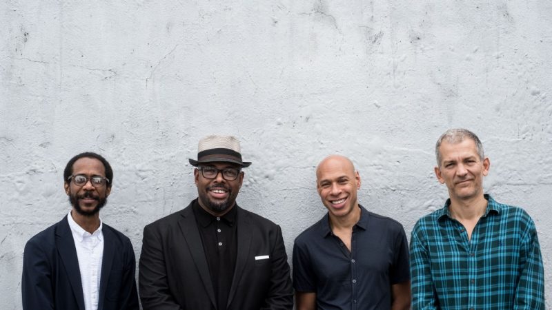 Musicians Joshua Redman, Brad Mehldau, Christians McBride, and Brian Blade pose in front of a stucco wall. The group consists of two Black men, one light skinned Black man, and a white man.