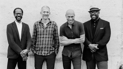  Musicians Joshua Redman, Brad Mehldau, Christians McBride, and Brian Blade pose in front of a stucco wall in this black and white image.