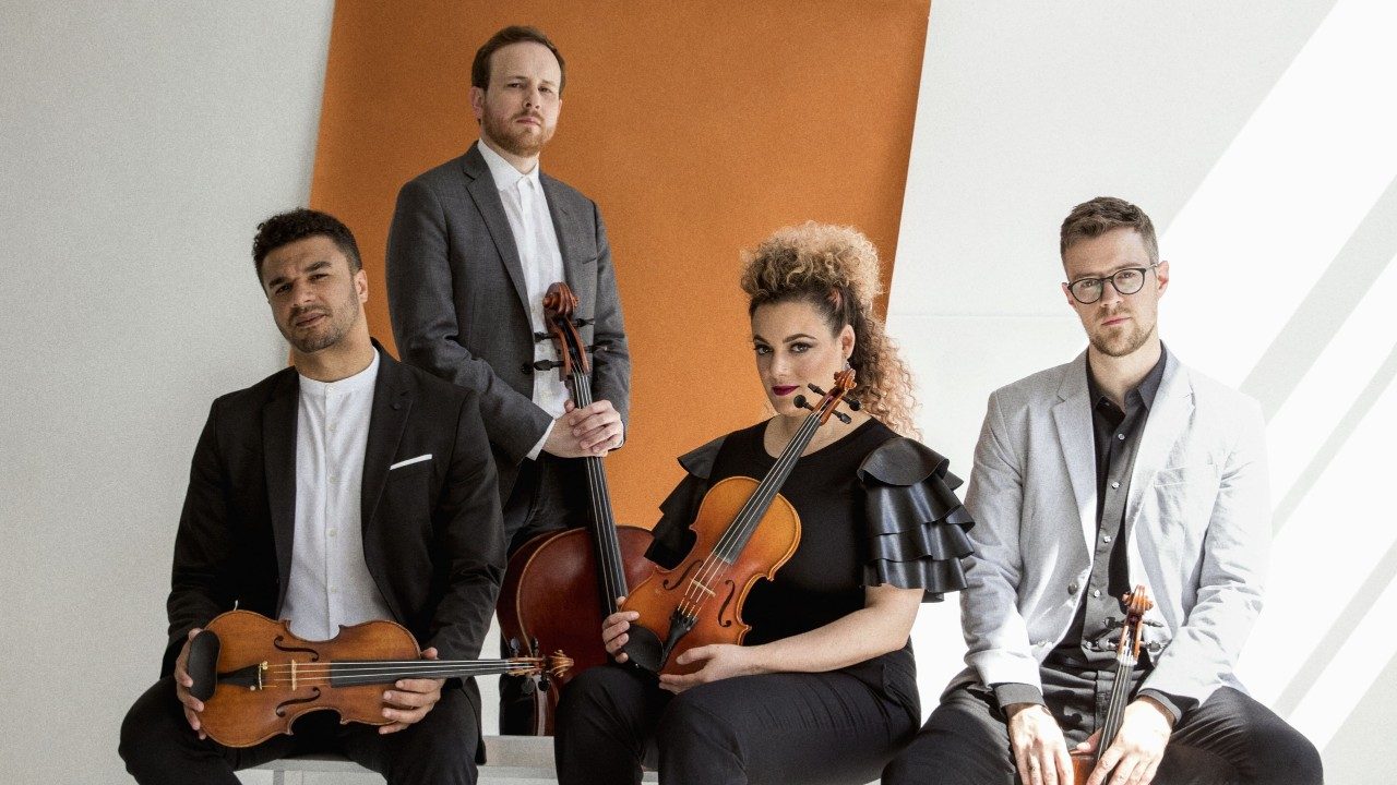  The members of PUBLIQuartet stand and sit in a photo studio holding their instruments. The room is painted white and a terracotta-colored roll of butcher paper hangs down the wall behind them. 
